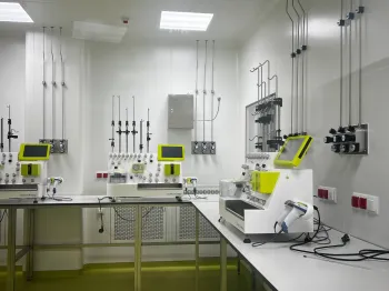 Laboratorium z białymi ścianami, zieloną podłogą, stołami i sprzętem laboratoryjnym.