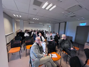 Grupa osób siedzących w sali konferencyjnej, niektórzy z notatkami, patrzą w stronę prezentacji.