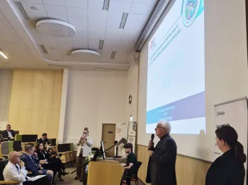 Sala konferencyjna, mężczyzna przemawia przy mównicy, publiczność siedzi, na ekranie prezentacja.