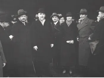 Grupa elegancko ubranych osób w płaszczach i kapeluszach, stojących na zewnątrz.