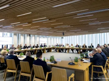 Osoby siedzące przy dużym, owalnym stole w sali konferencyjnej, na stole butelki wody.