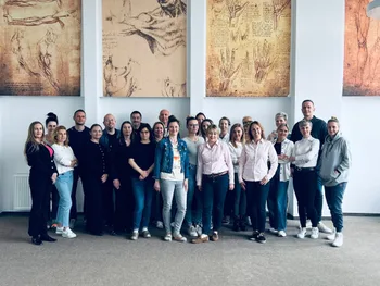 Grupa osób stoi w pomieszczeniu, w tle reprodukcje rysunków anatomicznych.