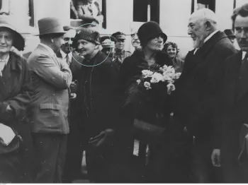 Grupa ludzi na peronie dworca, kobieta z kwiatami, mężczyźni w kapeluszach.
