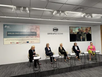 Pięć osób siedzi na scenie w sali konferencyjnej, z tyłu ekran z uczestnikami online.
