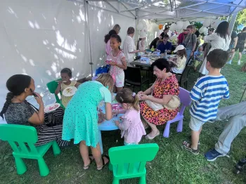Dzieci i dorośli przy stolikach pod namiotem na pikniku, na trawie, w otoczeniu drzew.