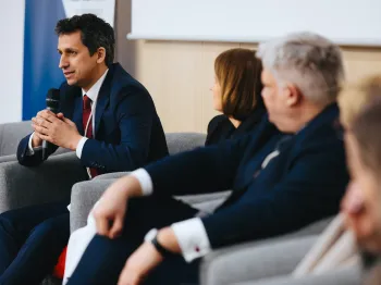 Mężczyzna w garniturze mówi do mikrofonu, siedząc obok innych osób na konferencji.