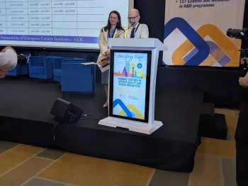 Dwie osoby stoją przy mównicy na scenie konferencyjnej, w tle ekran z prezentacją.