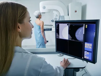 Kobieta w białym fartuchu patrzy na monitor z obrazami mammograficznymi, w tle badanie mammograficzne.