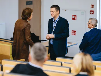 Trzy osoby rozmawiają w sali konferencyjnej, siedzenia w tle.