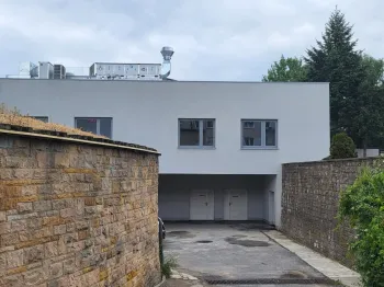 Budynek z białą fasadą, trzema oknami, otoczony murami z kamienia i zielenią.