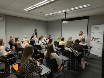 Grupa osób siedzi w sali konferencyjnej, jedna osoba stoi przy ekranie z logo Narodowego Instytutu Onkologii.