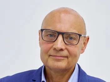 Mężczyzna w okularach i niebieskiej marynarce, patrzy wprost, jasne tło