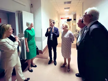 Grupa osób w korytarzu, rozmawiają, jedna osoba gestykuluje. W tle drzwi z napisem 