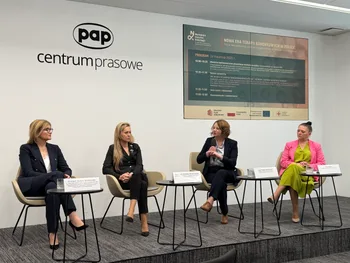 Cztery osoby siedzą na krzesłach w sali konferencyjnej, za nimi plakat z programem wydarzenia.